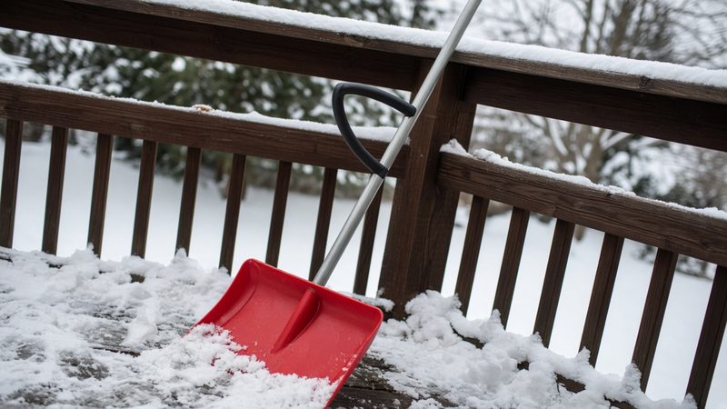 A red snow shovel with a second curved handle leaning on a snowy deck railing, in Detroit, Michigan